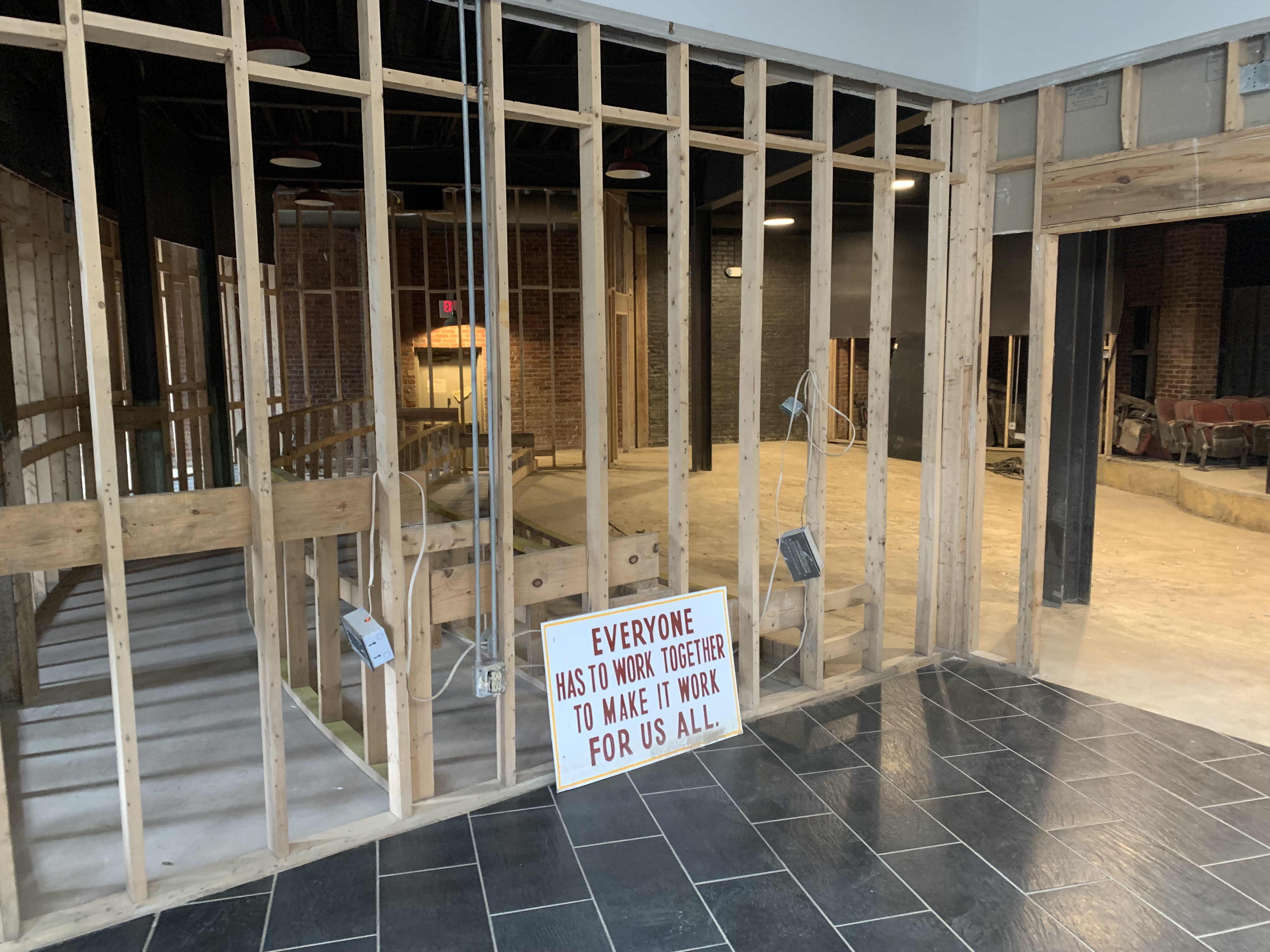 A photo of Appalshop's Theater space post-flood, structurally gutted with the walls and seats removed. Leaning against a post is a sign that reads "EVERYONE HAS TO WORK TOGETHER TO MAKE IT WORK FOR US ALL"