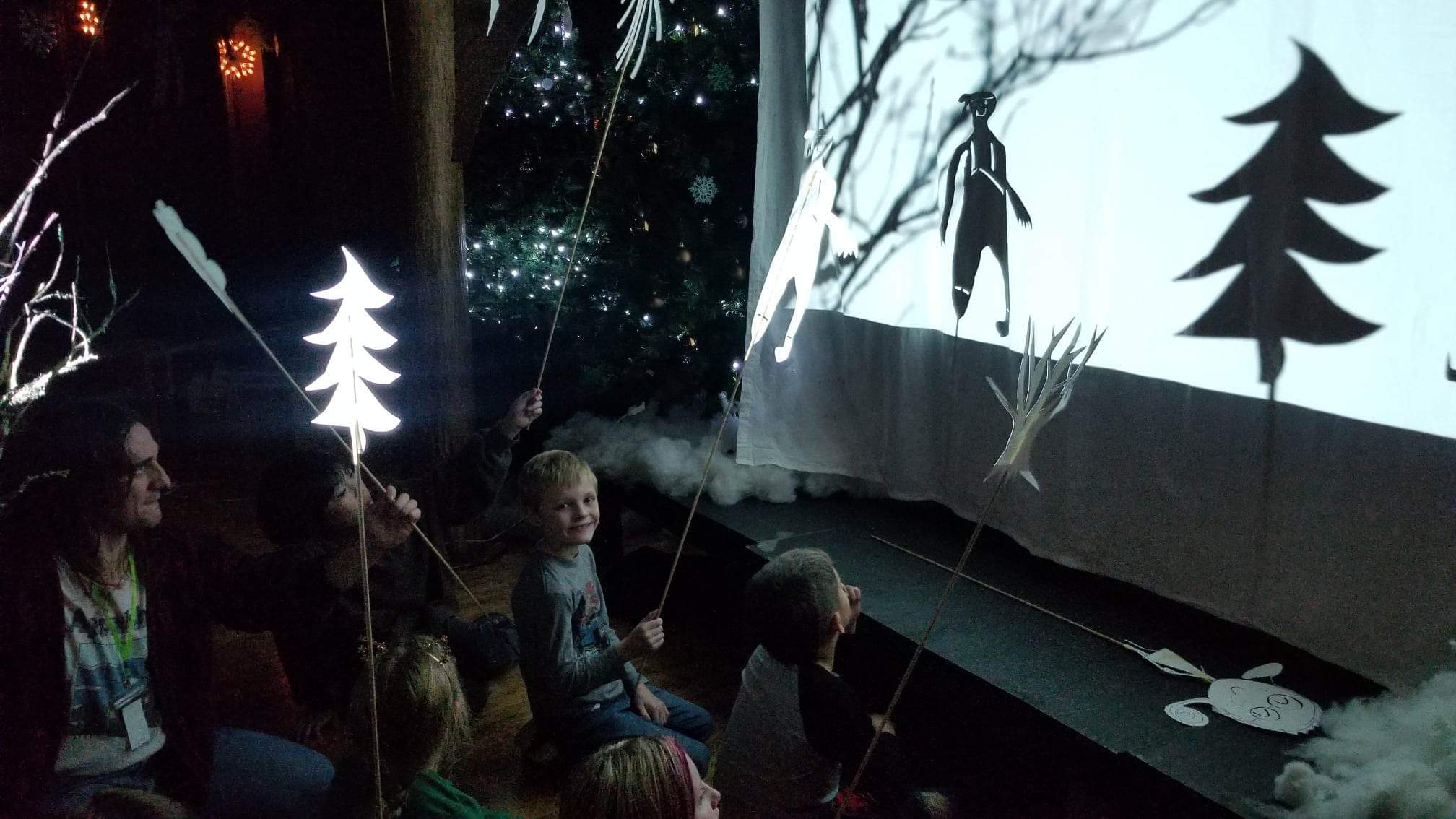 A photo of Tommy Anderson and a group of children crouching behind a projection screen, holding shadow puppets, performing a play. 